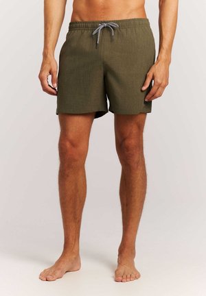 Homme portant un short de bain vert olive avec un cordon noir et blanc, debout pieds nus devant un fond clair uni.