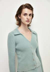 Ljusgrön cardigan gjord av mjukt, fluffigt material. Har en V-hals, långa ärmar och fem glänsande knappar fram. Texturerad stickning.