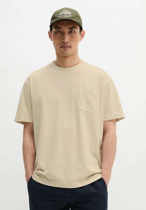 Man wearing beige short-sleeve T-shirt with breast pocket and olive green cap with white logo, standing with hands in navy pants pockets.