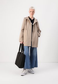 Casaco capuz bege com fecho éclair, combinado com uma camisa preta, jeans de perna larga e sapatilhas brancas de cano alto. Inclui uma mala tote preta.