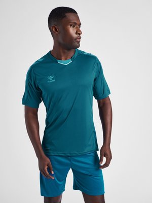 Mann trägt ein türkisfarbenes Sportshirt und Shorts, schaut zur Seite und steht vor einem schlichten hellgrauen Hintergrund.