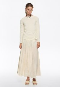 Maglietta a maniche lunghe color crema con coulisse laterale, abbinata a una gonna plissettata alla caviglia in una tonalità di crema più chiara e sandali beige semplici.
