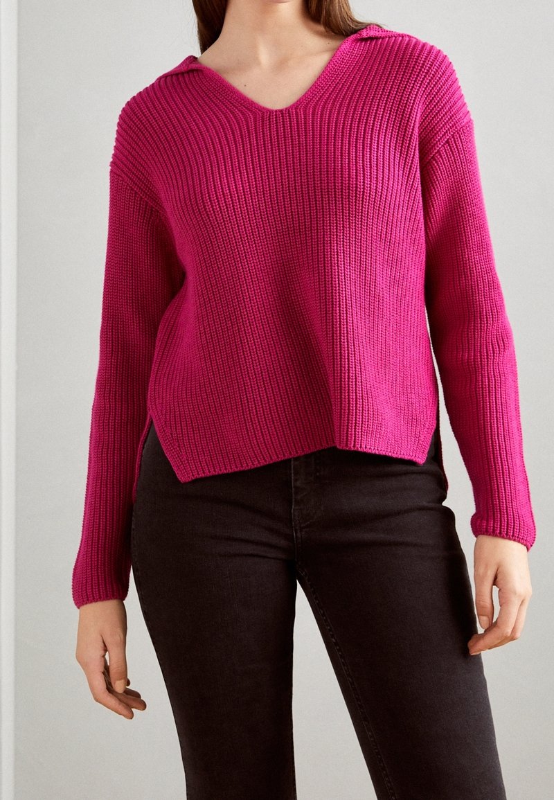 Femme portant un pull en maille rose vif à col en V, associé à un pantalon noir ajusté, debout devant un fond neutre.
