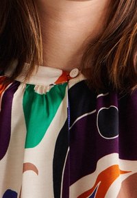 Färgglad blus med en blandning av gröna, lila och orangea former, med en samlad halsringning och en enda knappknäppning vid kragen.
