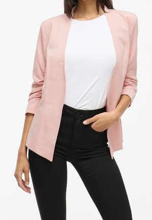 Mujer que lleva una blazer rosa claro con las mangas remangadas, top blanco y pantalones negros de talle alto, mano apoyada en el bolsillo.