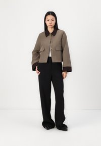 Brun jakke med cropped stil, store knapper og lommer, med en kontrasterende mørk krage. Brukt med svarte vidt utslåtte bukser og blanke sko.