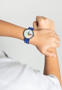 Montre pour enfants colorée avec un bracelet en caoutchouc bleu, un boîtier rond en argent et un cadran blanc affichant des chiffres en gras et une aiguille des heures colorée.