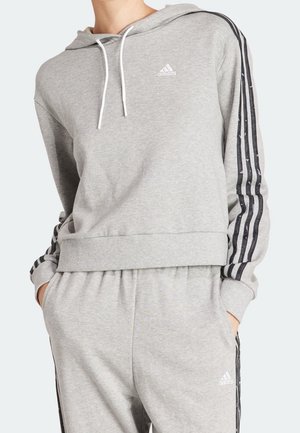 Osoba ubrana w szarą bluzę z kapturem Adidas i pasujące spodnie dresowe z czarnymi paskami na rękawach i nogawkach, ręce w kieszeniach.