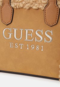 Tan schoudertas met een textuur, fluffy bovenkant, het gegraveerde "GUESS EST 1981" logo en gladde, contrasterende bruine leren accenten.