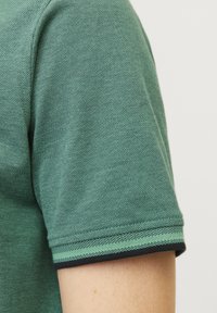 Gros plan sur une chemise verte à manches courtes avec tissu texturé et un ourlet rayé sur la manche portée sur un bras nu.