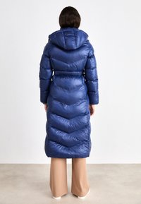 Duga puffer jakna tamnoplave boje, s uzorkom u obliku chevrona, kapuljačom i sužavajućim strukom, izrađena od sjajnog kviltanog materijala.