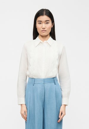 Νεαρή γυναίκα με ίσια σκούρα μαλλιά, ντυμένη με λευκό μπλουζάκι μακρυμάνικο με δαντελένιες λεπτομέρειες και ψηλόμεσα ανοιχτό μπλε παντελόνια.