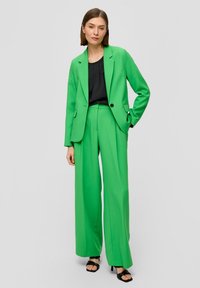 Groen op maat gemaakt pak met brede pijpen, met een blazer met één knoop en voorzakken. Draagt een zwart topje en is gecombineerd met zwarte hakken.