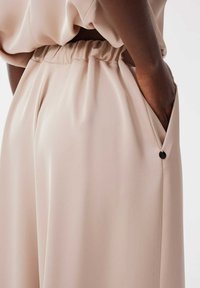 Beige, hoge taillebroek met een elastische tailleband, geplooid ontwerp, zijzakken en een gladde, lichte textuur. Zwarte knoopaccent.
