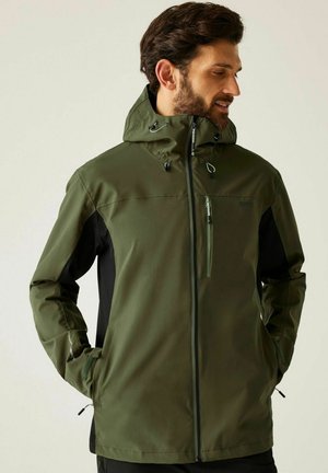 Hombre con chaqueta verde oliva y negra con capucha, de pie con las manos en los bolsillos, mirando hacia un lado contra un fondo liso.