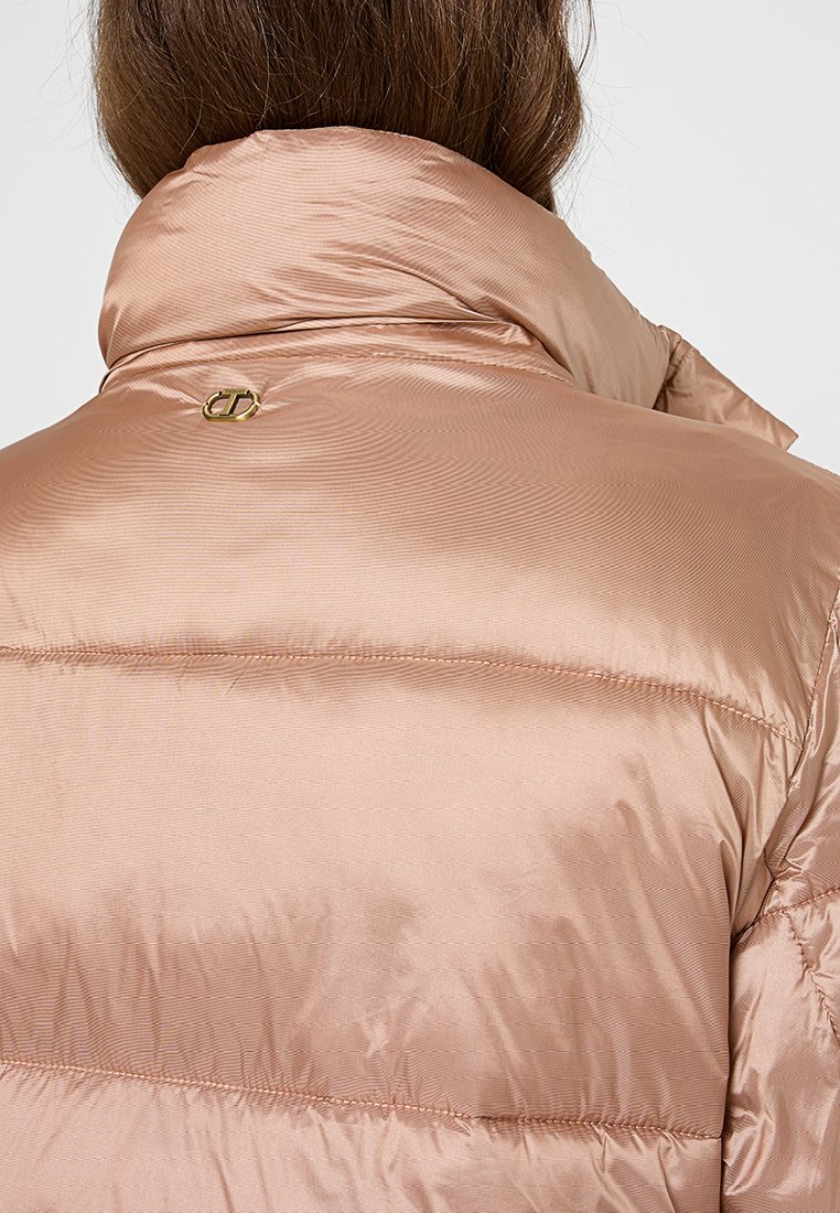 Padded jacket w jasnoróżowym satynowym materiale, z wysokim kołnierzem i subtelnym połyskiem. Posiada złoty akcent w postaci logo na plecach.
