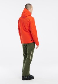 Heldere oranje waterdichte jas met capuchon, ritszakken, gecombineerd met groene broeken met zijritsen en zwarte skiboten.