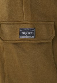 Khaki stoffbukse med en glatt tekstur og ribbet liv. Har en lommepatch og en svart etikett med teksten "PIER ONE" i hvite bokstaver.