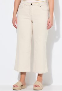 Pantalones vaqueros amplios recortados en color beige con cintura alta, bolsillos frontales y cierre de botón, combinados con sandalias de tira tejidas en tono marrón.