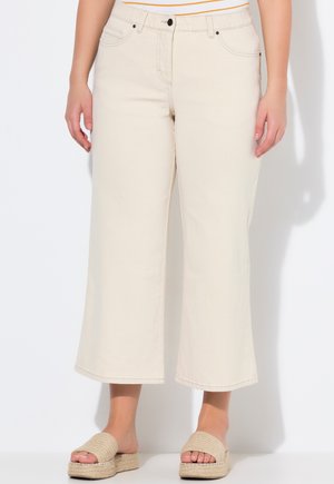 Jeans largo beige a vita alta, con tasche frontali e chiusura con bottone, abbinati a sandali a scivolo intrecciati color sabbia.