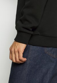 Mano derecha descansando a un lado, vistiendo una camiseta negra de manga larga y jeans de mezclilla azules con costuras de bolsillo visibles.