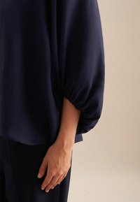 Blusa navy con maniche a palloncino, realizzata in tessuto leggero. Presenta un orlo arricciato sulle maniche e una vestibilità rilassata. Design minimal.