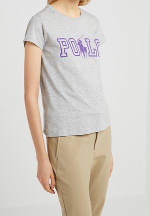 Kvinde iført en lysegrå t-shirt med korte ærmer, dekoreret med lilla "POLO" tekst og grafik af en polo spiller, kombineret med beige bukser mod en hvid baggrund.