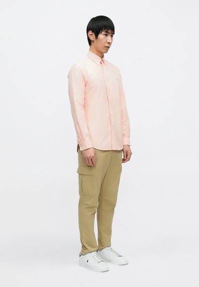 Ροζ ανοιχτό χρώμα, μακρυμάνικο πουκάμισο με κουμπιά, συνδυασμένο με μπεζ παντελόνια cargo και λευκά αθλητικά παπούτσια, με χαλαρή εφαρμογή και διακριτικές λεπτομέρειες στις τσέπες.