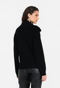 Maglione nero a maglia con colletto alto, maniche lunghe e texture a coste. Il modello lo indossa con pantaloni in pelle nera, evidenziando un design minimalista.