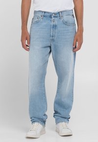 Framifrån vy av person som bär ljusblå raka jeans, vita sneakers och en vit inskjuten t-shirt, stående mot en enkel bakgrund.