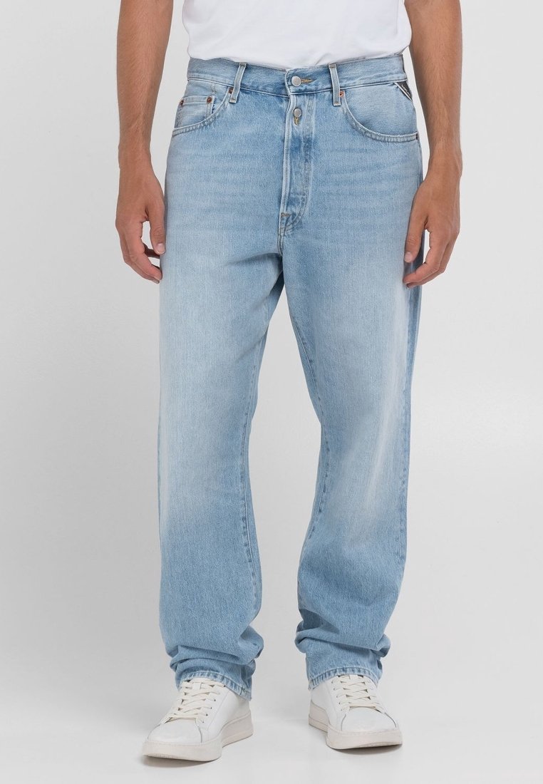 Framifrån vy av person som bär ljusblå raka jeans, vita sneakers och en vit inskjuten t-shirt, stående mot en enkel bakgrund.