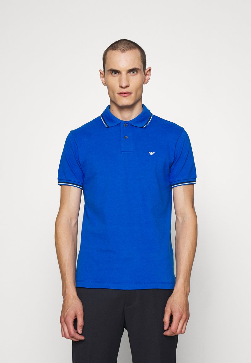 Μπλε πόλο μπλούζα με γιακά, με διπλές λευκές και μπλε ρίγες στον γιακά και τα μανίκια, και ένα μικρό λογότυπο στο στήθος.