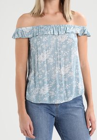 Svetle modrý top bez ramínek s řaseným výstřihem; má bílé květinové vzory a volný střih, doplněný modrými džíny.