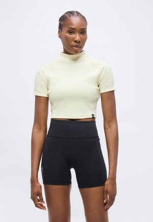 Kvinde iført en lysegul kortærmet cropped top og højttaljede sorte træningsshorts, stående mod en ensfarvet hvid baggrund.