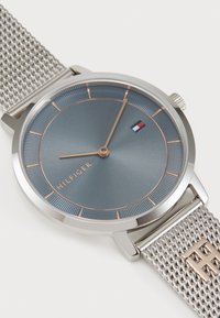 Sølv mesh-remmewatch med en rund, mørkeblå urskive, roseguld accenter og minimalistiske timemarkører. Har "HILFIGER" logo.