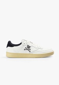 Zapatilla de cuero blanca con acentos negros, logo de calavera y huesos cruzados en el costado, suela de goma texturizada y puntera perforada para transpirabilidad.