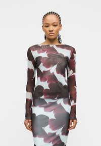 Γυναίκα με πλεξούδες που φοράει μακρυμάνικη μπλούζα και φούστα με μεγάλο μοτίβο μαργαρίτας σε μπορντό και λευκό χρώμα σε διάφανο ύφασμα.