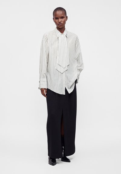 Personne portant une chemise blanche oversize à rayures verticales avec un col prolongé et une longue jupe noire avec une fente à l'avant.