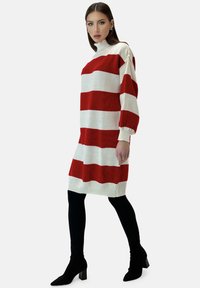 Rood-wit gestreepte trui-jurk met een hoge hals, voorzien van lange, geplooide mouwen en een gebreide textuur. Gedragen over zwarte legging en laarzen met hak.