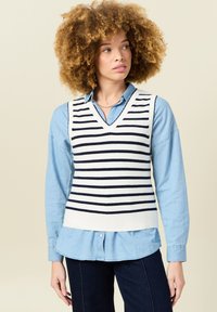 Navy- und weiß gestreifter Strickweste mit V-Ausschnitt, über einem hellblauen Hemd mit Knopfleiste getragen, kombiniert mit dunklen Jeans.