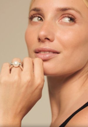 Mujer de piel clara y cabello rubio que lleva un anillo de perlas, con el puño cerca de la barbilla mientras mira hacia un lado.