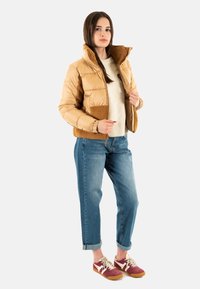 Vadderad beige kort jacka med bruna fleecekanter och ficka, bärs över en krämfärgad tröja, kombinerad med blå jeans och brunröda sneakers.