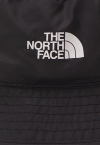 Fekete vödörsapka textúrált anyagból, fehér logóval, "THE NORTH FACE" felirattal, ívelt ívvel és vízszintes varrással.