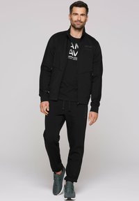 Zwarte sportjack met rits, bijpassend zwart t-shirt met witte tekst, en zwarte joggers. Draagt groene sneakers.