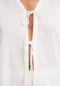 Blusa bianca in lino con scollatura a V, legata davanti con due asole in tessuto e un sottile patta; tessuto leggero e testurizzato con un aspetto semi-trasparente.