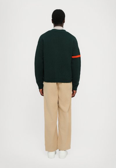 Suéter de punto verde con una franja naranja en la manga izquierda, combinado con pantalones de pierna ancha beige y zapatillas blancas.