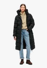 Mujer vestida con un abrigo de plumas negro largo, un suéter beige con cremallera, jeans celestes remangados y botas marrón con cordones, de pie con una mano en el bolsillo.