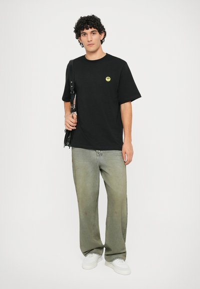 T-shirt en coton noir avec un petit logo jaune fluo, associé à un jean vert clair et des baskets blanches. Le modèle tient un sac noir.