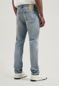 Man in lichtblauwe vervaagde jeans en witte sneakers, staande met zijn rug naar de camera op een effen achtergrond.