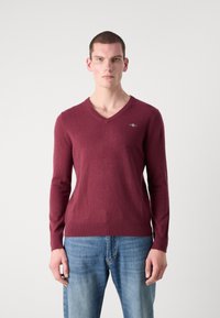Pulover vișiniu cu decolteu în V și mâneci lungi, fabricat dintr-un material moale. Prezintă un mic logo pe piept și un design ajustat la talie.